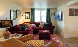 Una sala de estar con un sofá rojo, un sillón de cuero marrón, un reposapiés estampado, una lámpara decorativa y una alfombra colorida.