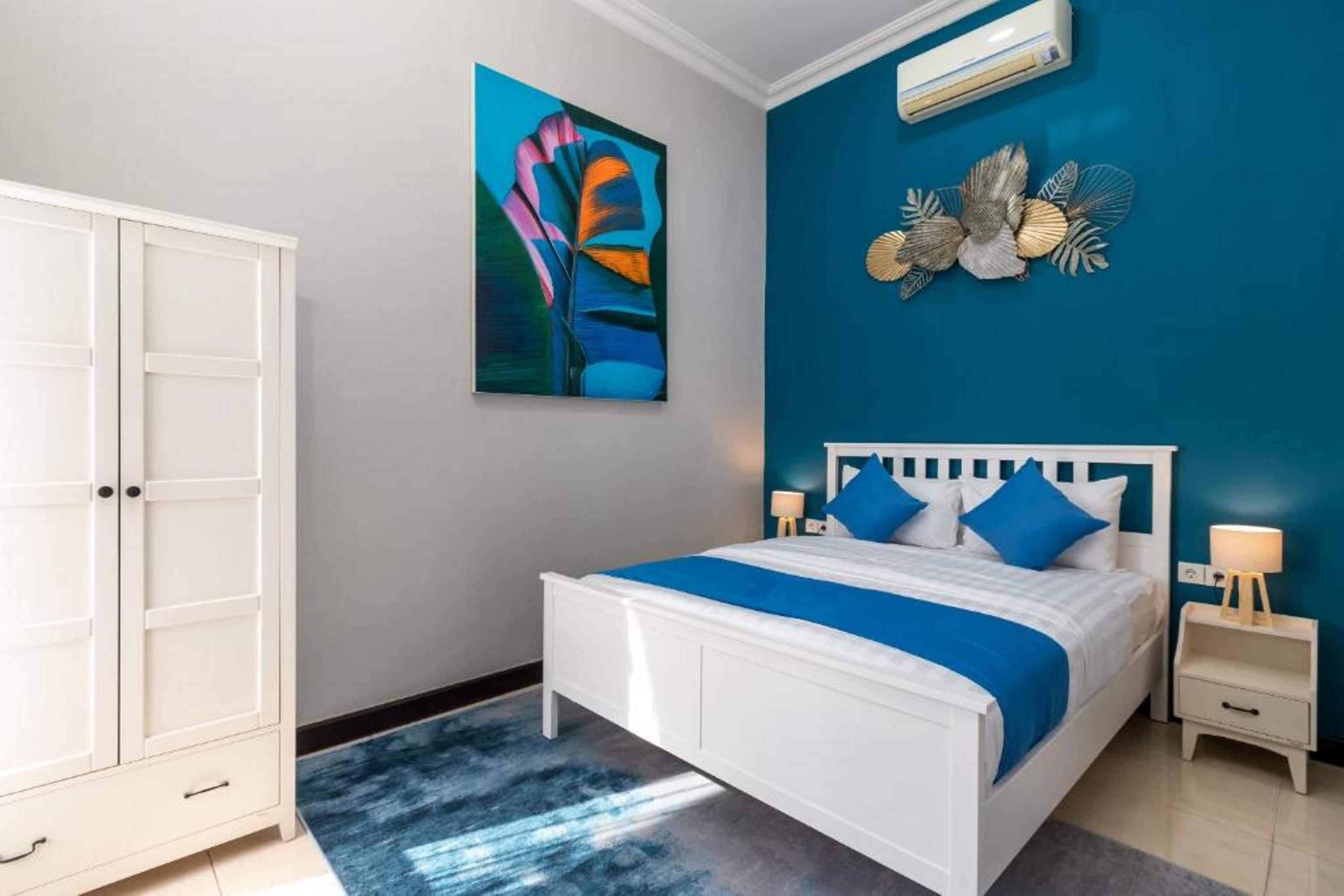 Une chambre à coucher avec un lit blanc, une literie bleue, une armoire blanche et un tableau accroché au mur.