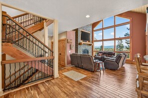 A spacious living area with a wooden staircase, a fireplace, and a large window offering a view of the outdoors. - West Cobb- Private- sleeps 16- hot tub- petsOK (McCall)