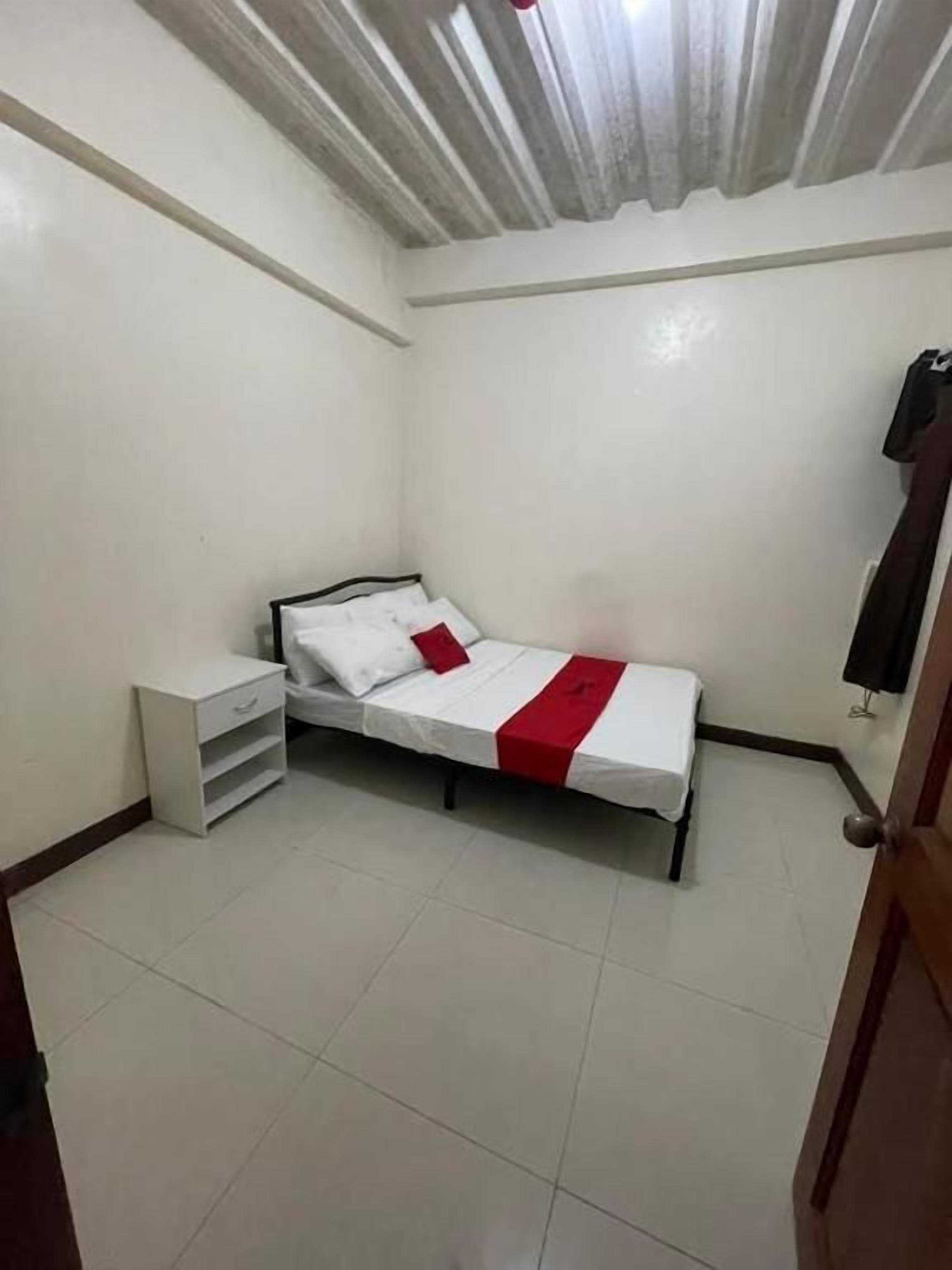 Una habitación pequeña con cama individual, cubrecama blanco, almohada decorativa roja, mesita de noche blanca y una puerta a la derecha.