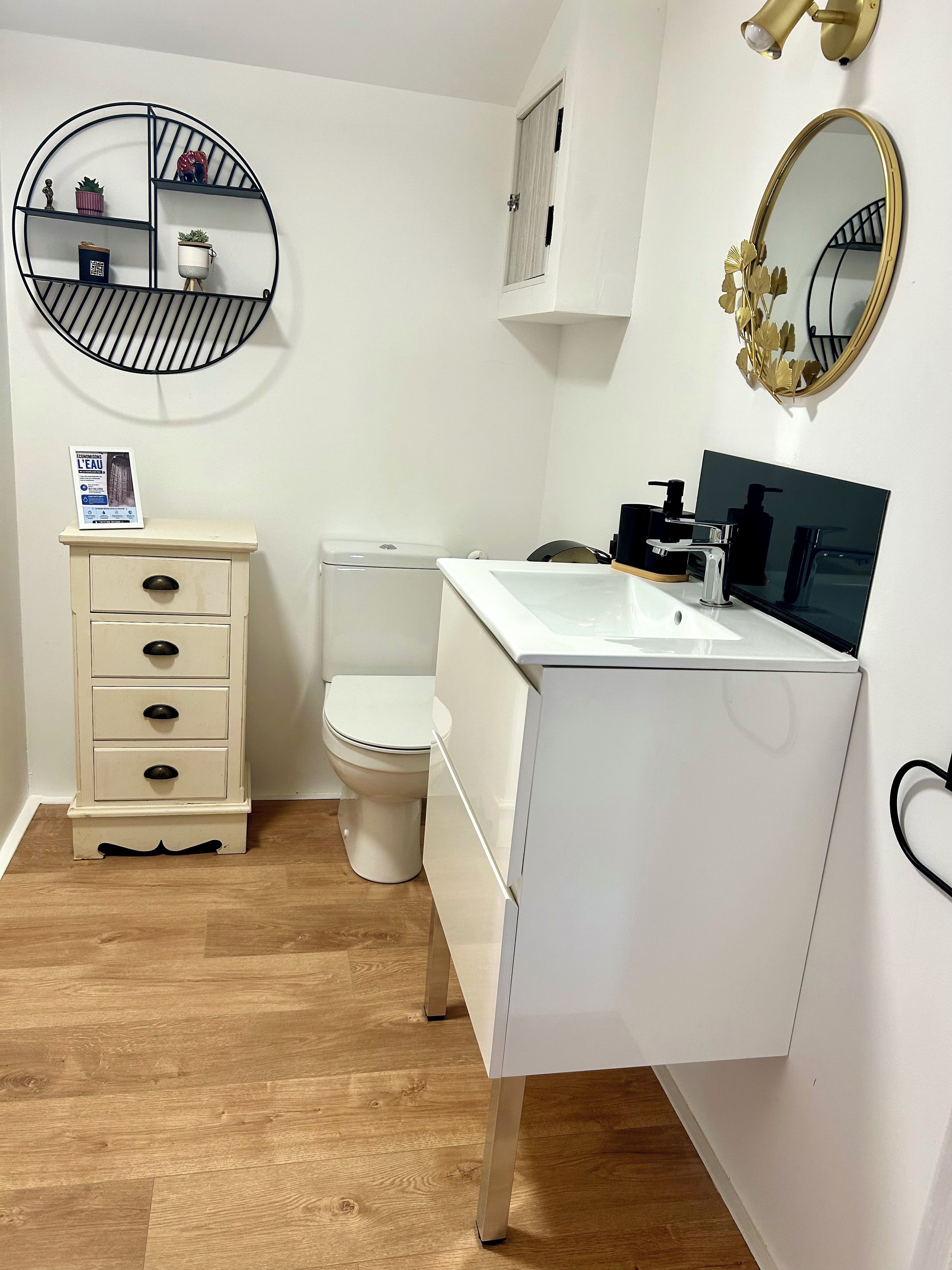 A bathroom with a toilet, a sink with a mirror, a round shelf with decorative items, and a cabinet with drawers.