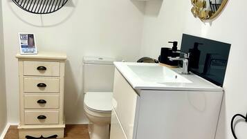 A bathroom with a toilet, a sink with a mirror, a round shelf with decorative items, and a cabinet with drawers.