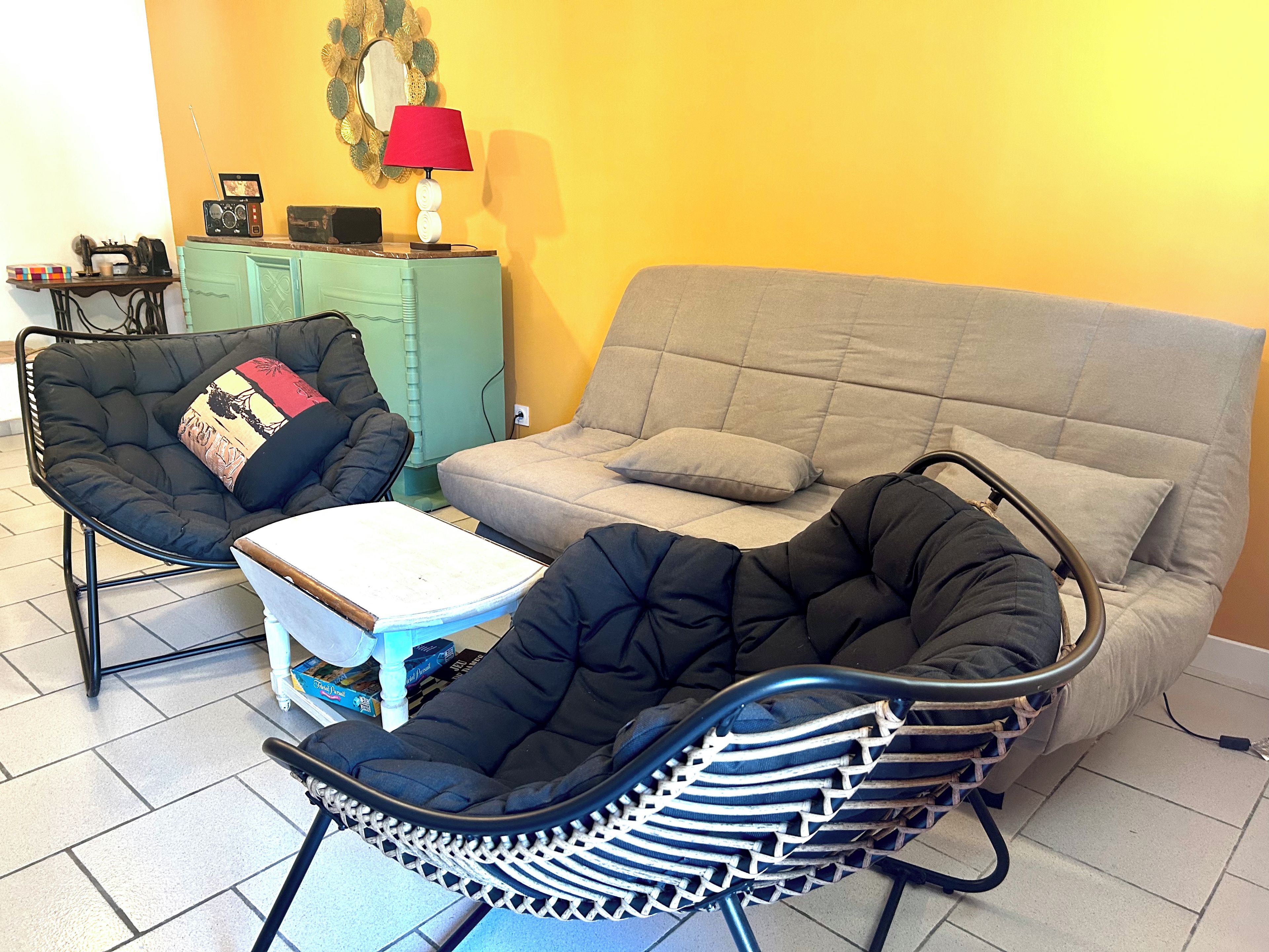 Une pièce avec un canapé gris, un fauteuil noir et une chaise en osier, accompagnée d’une table blanche.