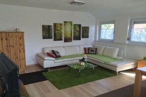 Ein modernes Wohnzimmer mit einem weißen Sofa, einem Glastisch und einem grünen Teppich. Vier gerahmte Bilder hängen an der Wand, und ein Holzschrank ist zu sehen.