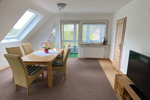 A room with a dining table, chairs, a television, and a window with a view of greenery. - Holiday apartment with 1 bedroom (Freudenstadt)