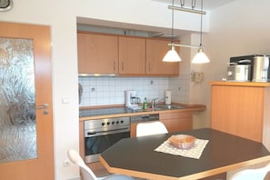 A compact kitchen with wooden cabinets, a stove, a sink, and a dining table with a loaf of bread. - Holiday apartment with 2 bedrooms (Seebad Heringsdorf)