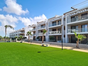 A modern multi-story residential building with balconies, a well-maintained lawn, and palm trees. - Ground Floor Apartment Overlooking the Pool (Los Alcazares)
