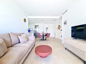 A modern living room with a sofa, a pink ottoman, a dining area with chairs, a kitchen with a fridge, and a TV on a stand. - Ground Floor Apartment Overlooking the Pool (Los Alcazares)