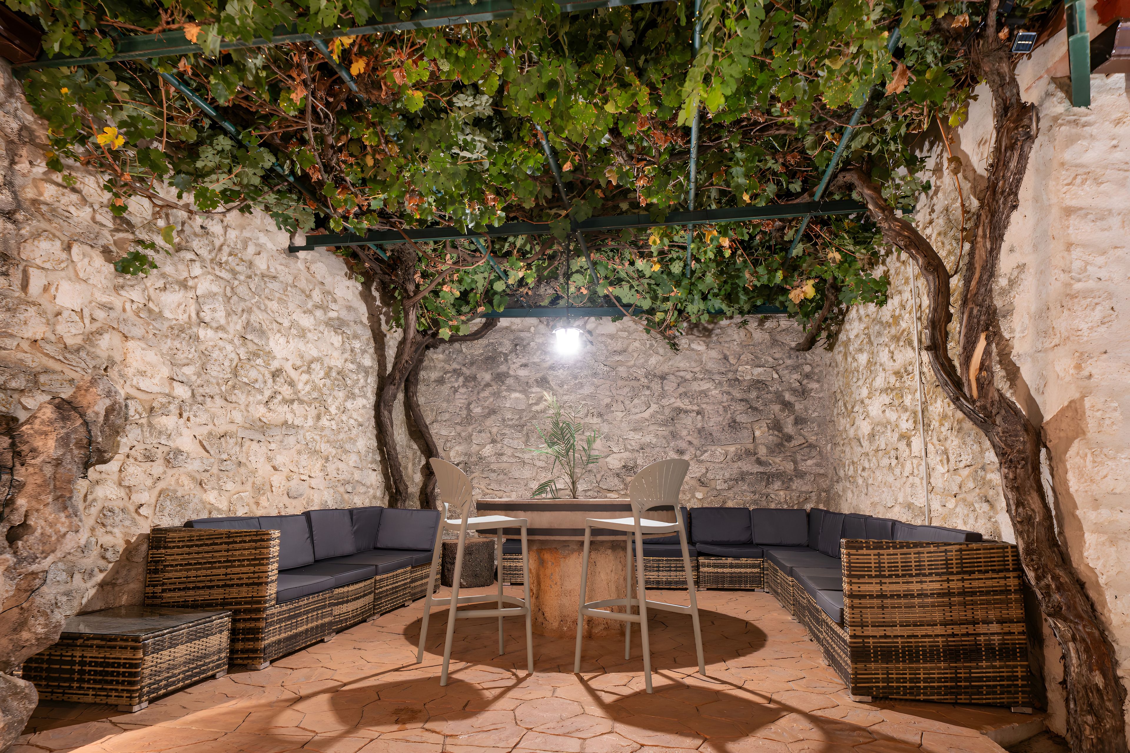 Un espace extérieur aménagé avec des murs en pierre, comprenant une table et des chaises, entouré de meubles en osier et d’une plante grimpante suspendue.