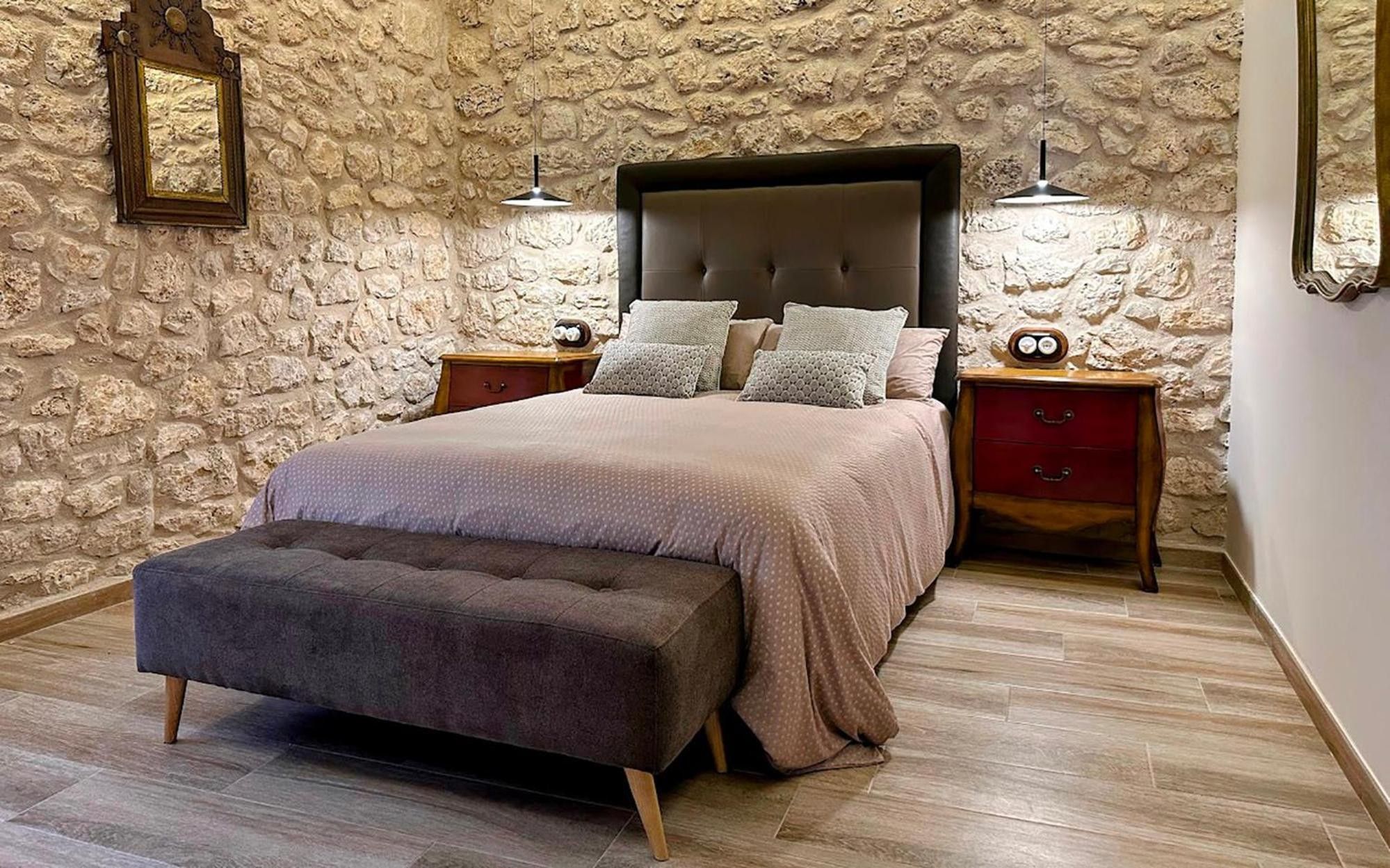A bedroom with a stone wall, a bed with a tufted headboard, two bedside tables with lamps, and a bench at the foot of the bed.