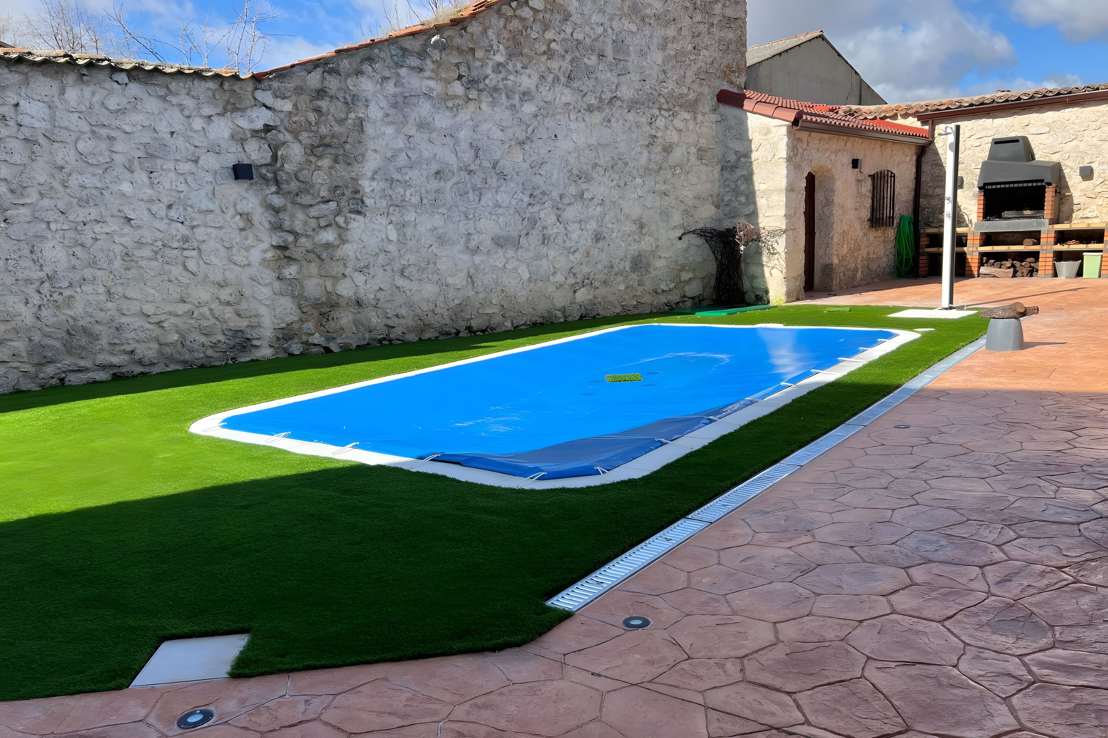 A swimming pool with artificial grass, a stone wall, and a tiled patio.