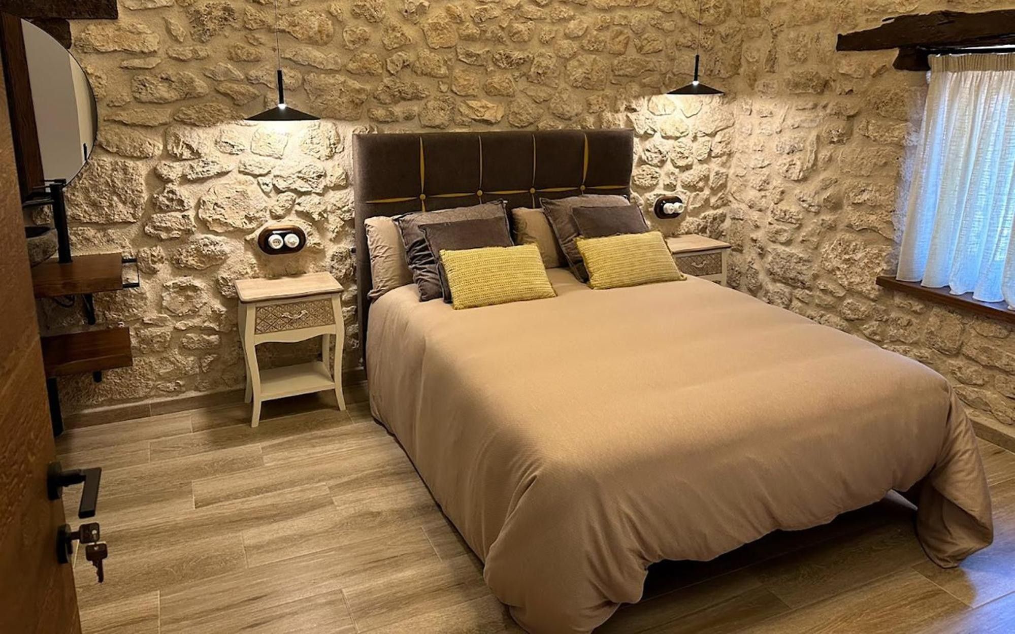 Un dormitorio con pared de piedra, una cama con varias almohadas, mesitas de noche y una ventana con cortinas.