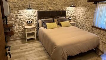 Un dormitorio con pared de piedra, una cama con varias almohadas, mesitas de noche y una ventana con cortinas.
