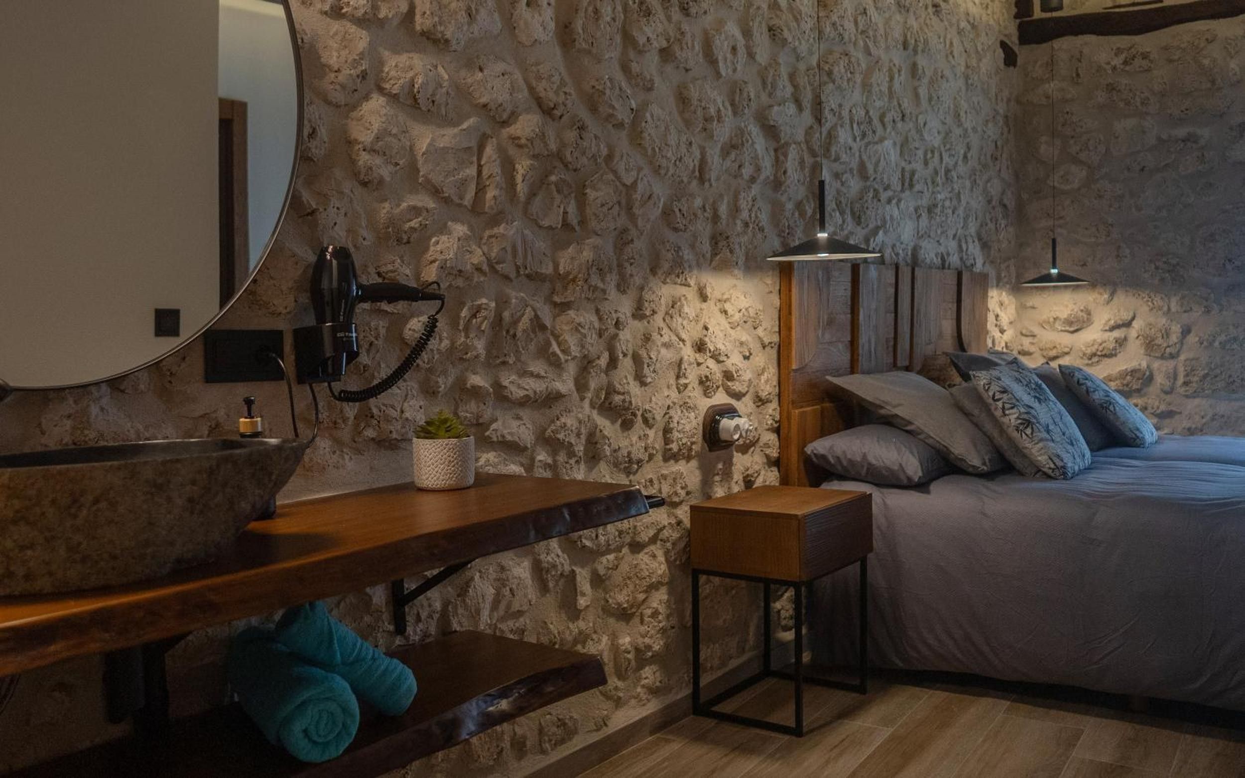 Un dormitorio con pared de piedra, una cama con almohadas, una mesita de noche, una lámpara y un espejo.