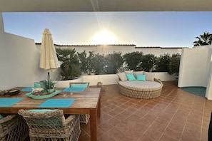 Eine Terrasse im Freien mit einer runden Sitzecke, einem Holztisch mit Stühlen und einem Sonnenschirm.