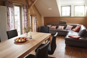A dining area with a wooden table, chairs, and a fruit bowl. There is a sofa with cushions in the background. - Blue holiday apartment (Michendorf)