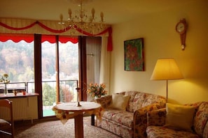 A living room with a sofa, a table, a chandelier, a clock, and a window with a view of trees. - Holiday apartment with 2 bedrooms (Bad Herrenalb)