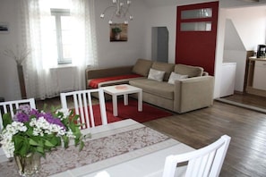 A living room with a sofa, a small table, and a dining table with chairs. There is a window with curtains, a plant, and a red door. - Holiday apartment with 2 bedrooms (Waren-Müritz)