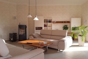 Ein Wohnzimmer mit einer Couch, einem Couchtisch und Pendelleuchten. Außerdem ist ein Bücherregal und ein Topf mit einer Pflanze zu sehen.
