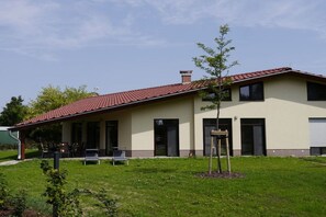 Een gebouw met twee verdiepingen, een rood pannendak, een overdekt terras met zitplaatsen buiten en een kleine boom in de voortuin.