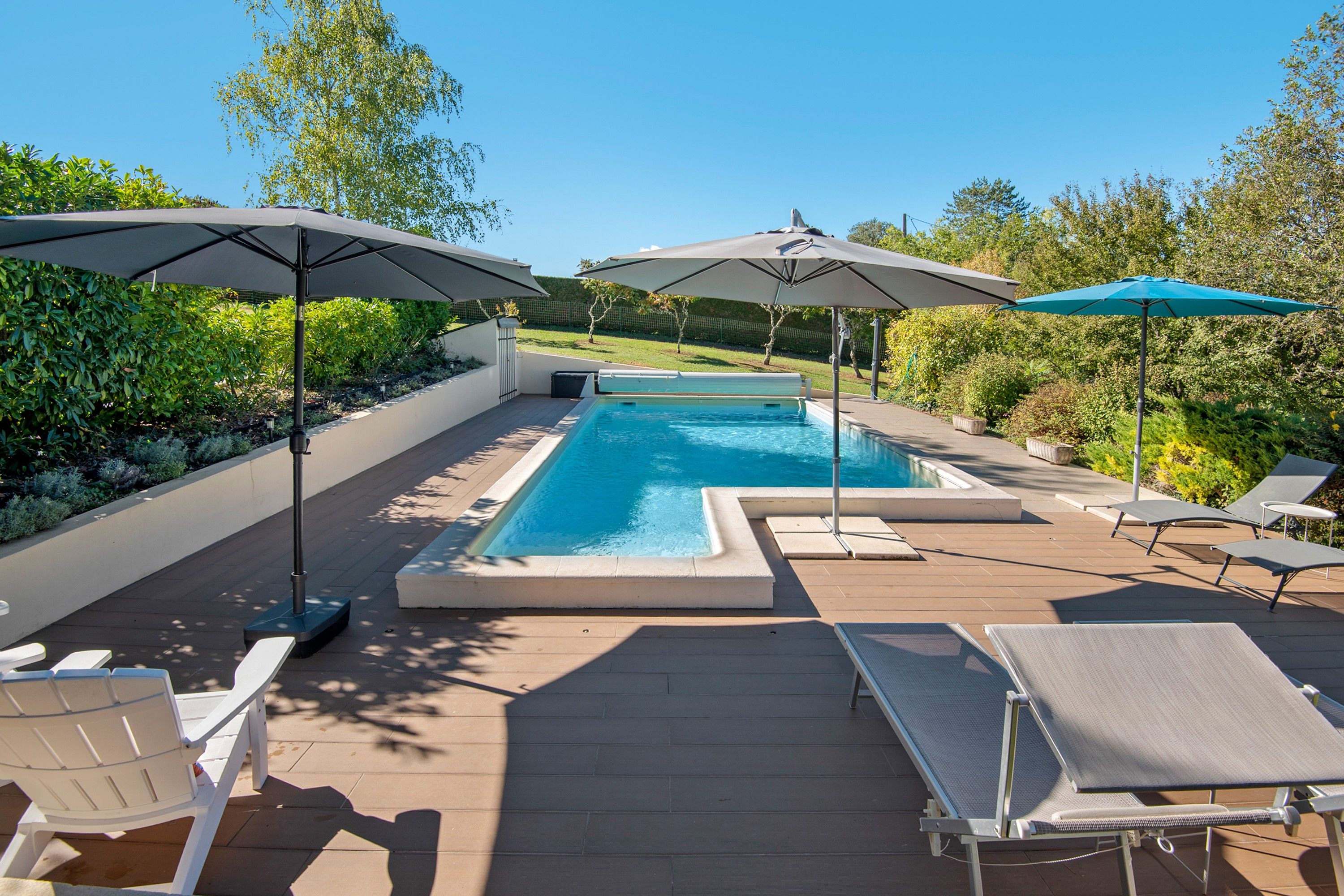 A rectangular swimming pool with a wooden deck, surrounded by lounge chairs and umbrellas.