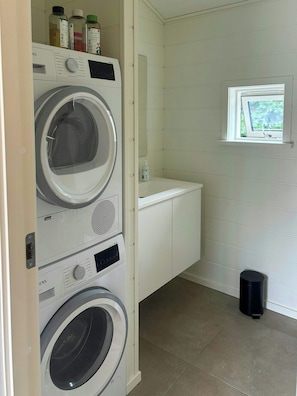 Een wasserette met een wasmachine en droger boven elkaar, een klein raam en een afvalbak.