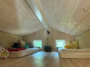A small attic room with wooden slats on the ceiling, two windows, and a mattress on the floor.