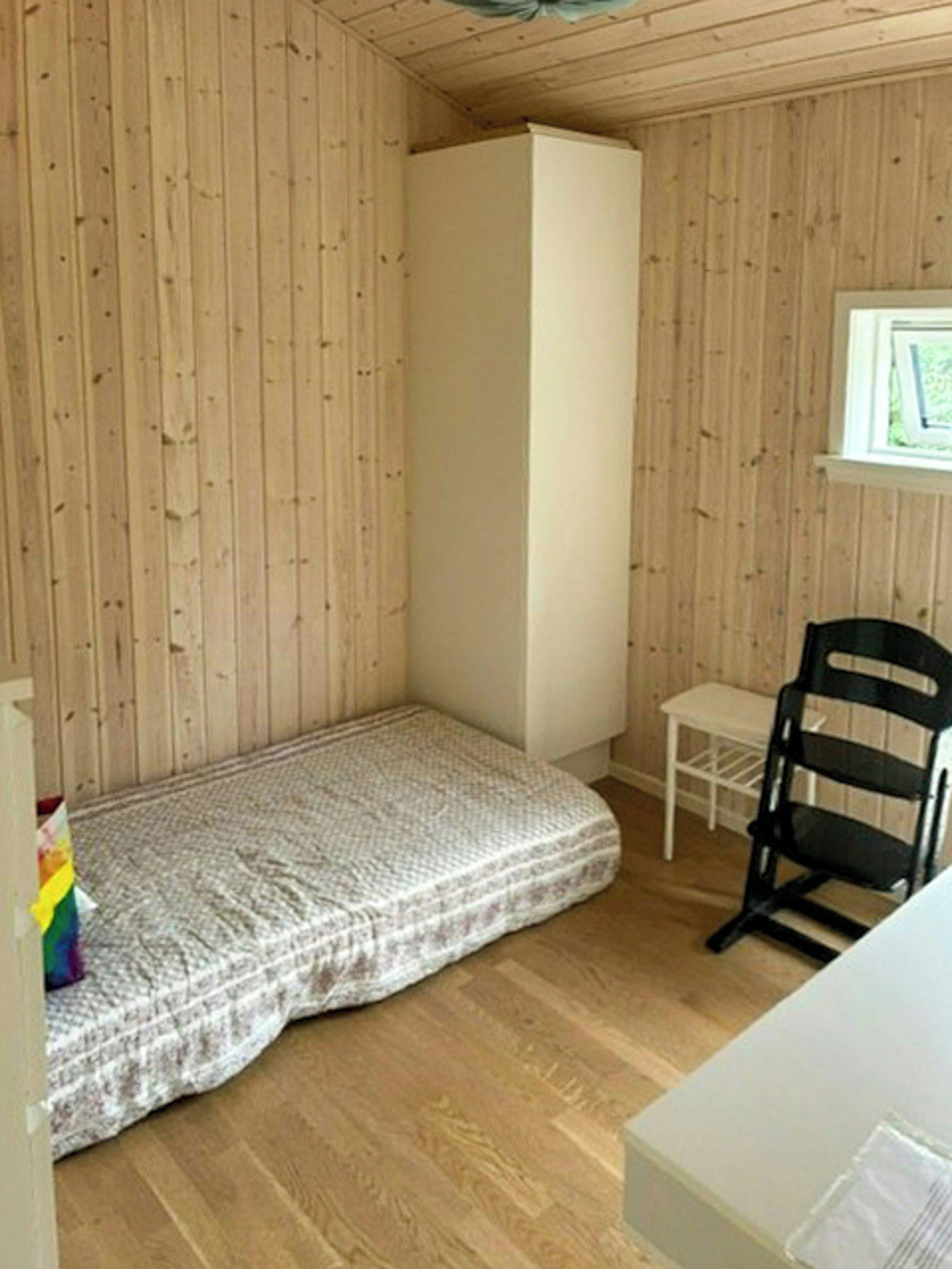 A small room with wooden walls, a bed, a chair, and a window.