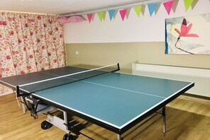 Ein Tischtennis-Tisch in einem Raum mit einem blumigen Vorhang, einem Heizkörper und einer Wand mit farbenfroher Wimpelgirlande.