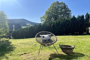 Un espacio al aire libre con césped, una silla de mimbre y una cesta, una casa al fondo, y un cielo despejado.