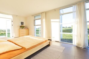 Ein Schlafzimmer mit einem großen Bett, einem Schminktisch und Blick auf eine grüne Landschaft durch große Fenster.