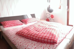 Ein Bett mit kariertem Überwurf und Kissen, ein Nachttisch mit Lampe sowie eine Wand mit Lichtschalter und Dekorationselement.