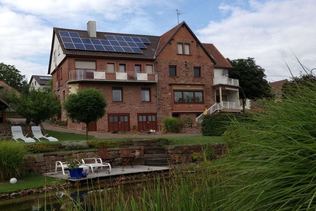 A two-story house with a solar panel roof, a small garden, and a pond.
