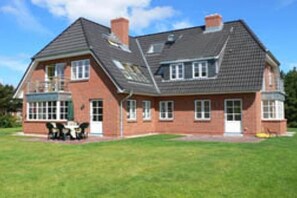 A two-story house with a brick exterior, a front porch with outdoor furniture, and a large lawn. - Holiday apartment with 2 bedrooms (Norddorf)