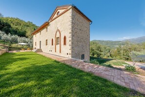 Ein Steinhaus mit einem geneigten Dach, einem gepflasterten Weg und einer Rasenfläche mit einer Bank.