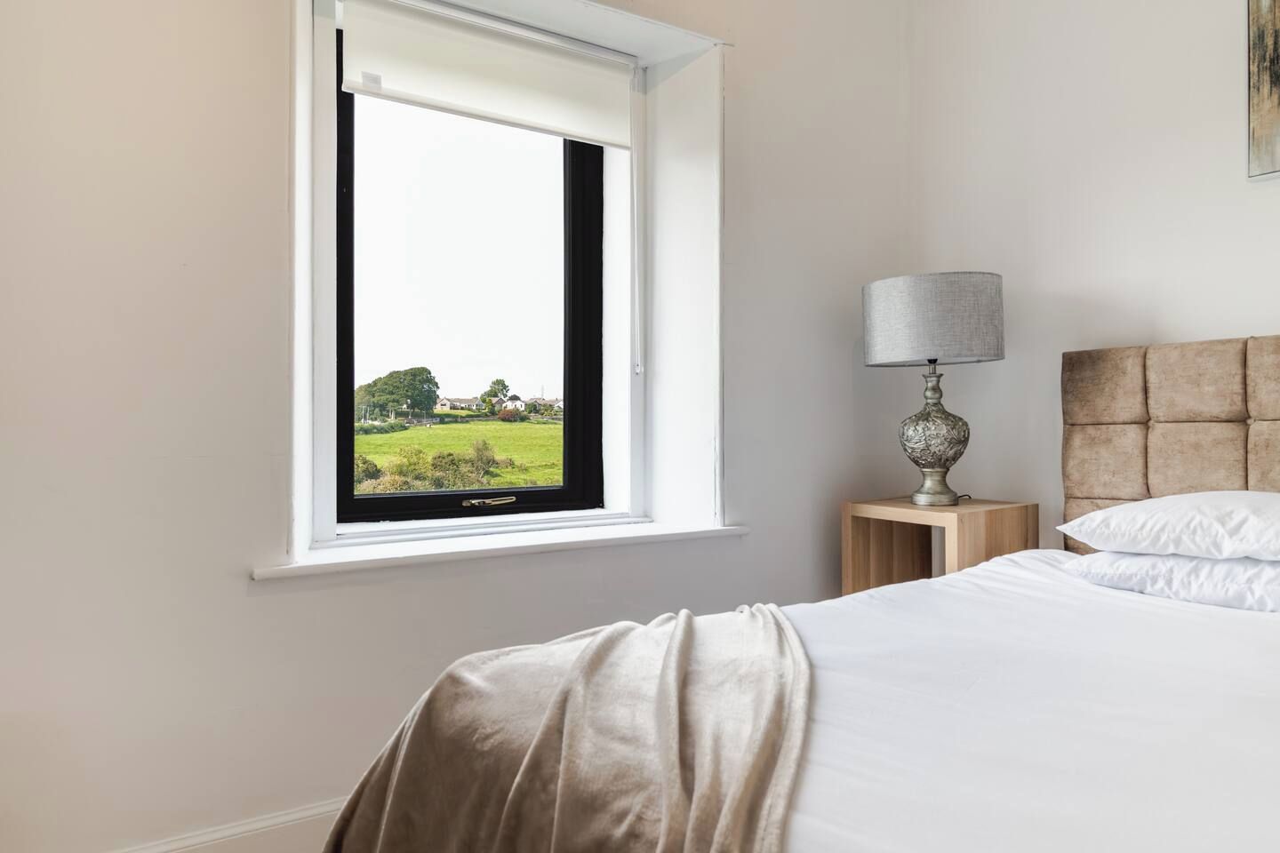 Une chambre à coucher avec un lit, une table de chevet avec une lampe, une fenêtre donnant sur un paysage verdoyant et une tête de lit.