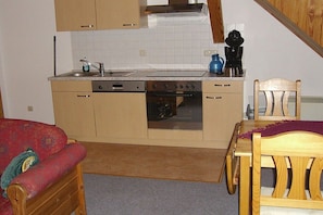 Eine kompakte Küche mit Herd, Backofen und Spüle. Im angrenzenden Wohnbereich befindet sich ein Esstisch mit Stühlen sowie eine Couch.