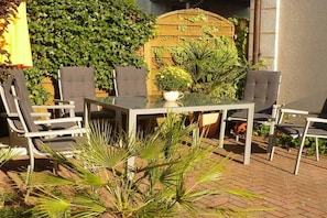 Eine Terrasse im Freien mit einem Glastisch und Stühlen, umgeben von Kübelpflanzen und einem Holzzaun.