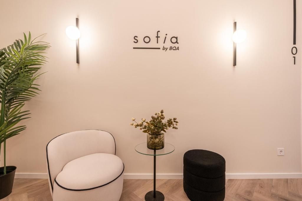 A room with a white chair, a black ottoman, a small table with a vase of flowers, and wall-mounted lights.