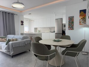 A modern living room with a grey sofa, a round dining table with four chairs, and a kitchen area with white cabinets and appliances. - Vacation Apartment (Vejer de la Frontera)