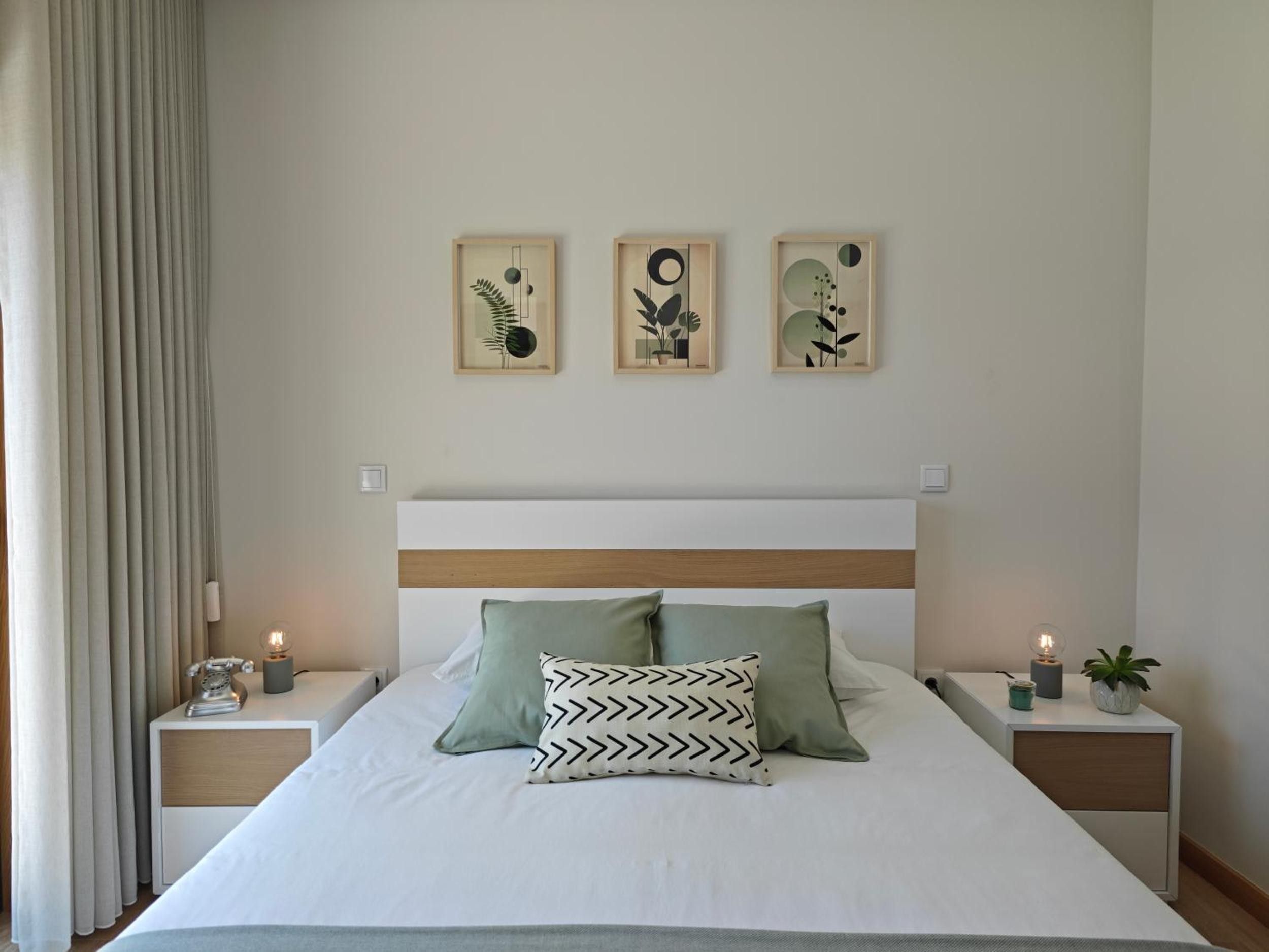 Een net opgemaakt bed met twee kussens, een nachtkastje met een lamp, en drie ingelijste kunstwerken aan de muur.