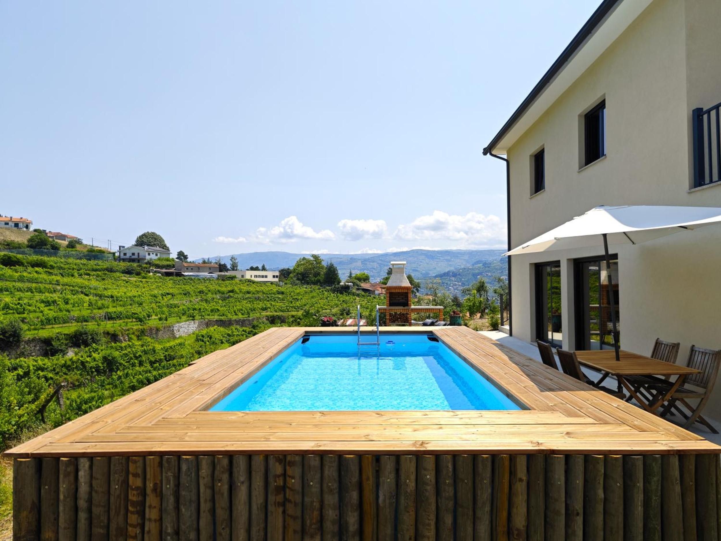 A wooden deck with a swimming pool, a brick barbecue, and a view of a vineyard and hills.