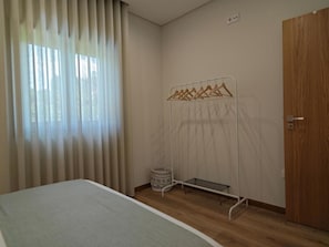 Un dormitorio con cama, una puerta de madera, una ventana con cortinas y un tendedero metálico.