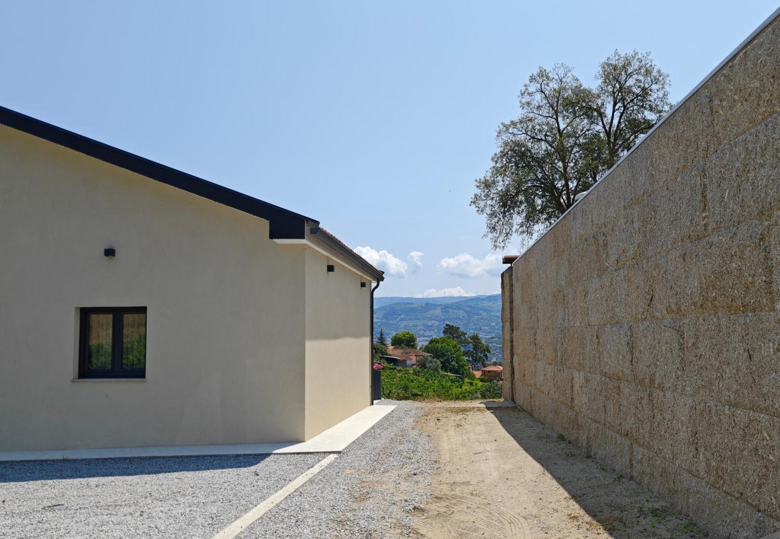 A building with a sloped roof and a small window, adjacent to a stone wall with a gravel path leading to a view of distant houses and mountains.