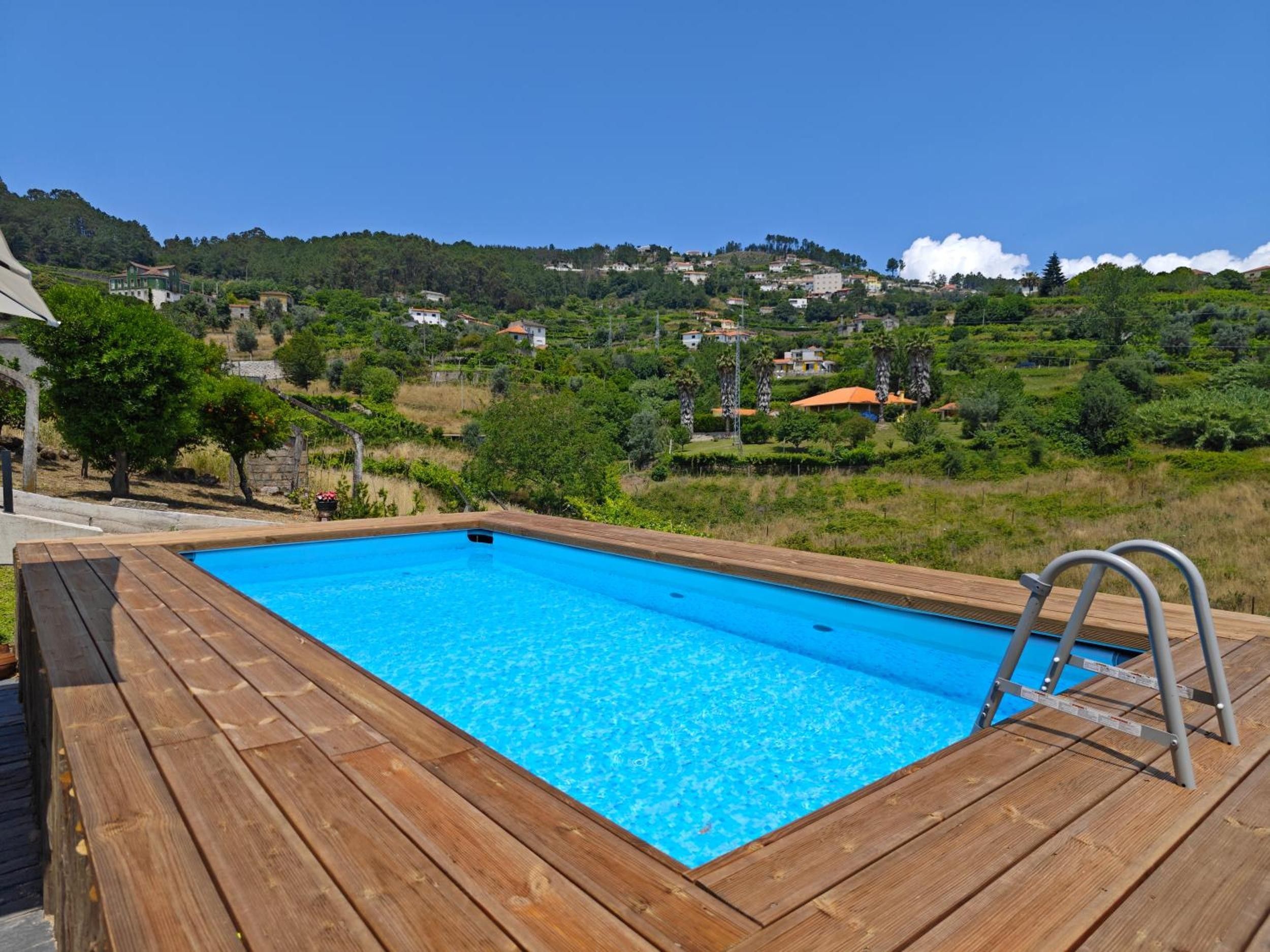 A wooden deck with a swimming pool, a ladder, and a view of a hillside with houses.