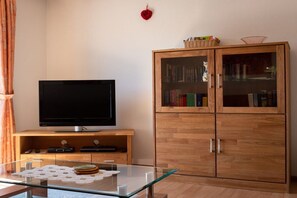 A living room with a flat-screen TV, a glass-top coffee table, a wooden cabinet with books, and a red decorative item on the wall. - Holiday apartment with 1 bedroom (Neunkirchen)
