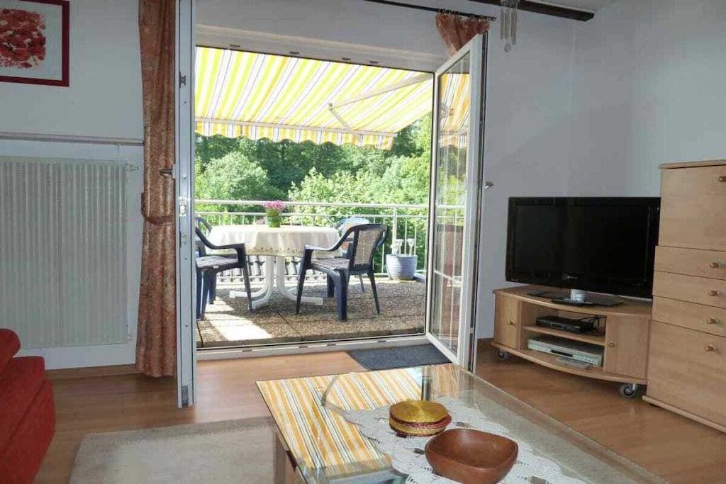 A living room with a TV, a sofa, a glass table with decorative items, and a sliding glass door leading to an outdoor area with a table and chairs.