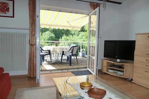 A living room with a TV, a sofa, a glass table with decorative items, and a sliding glass door leading to an outdoor area with a table and chairs. - Holiday apartment with 1 bedroom (Neunkirchen)