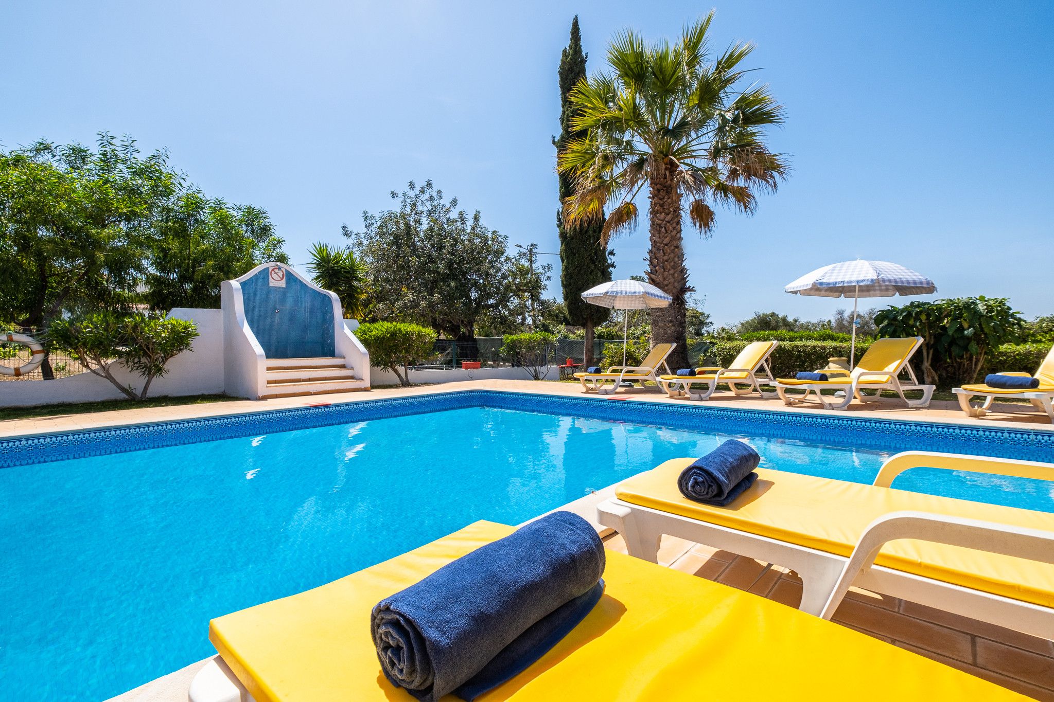 Une piscine entourée de chaises longues et de parasols, bordée de palmiers et surplombée d’un ciel dégagé.