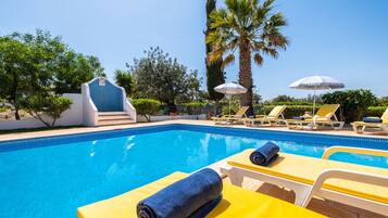 Une piscine entourée de chaises longues et de parasols, bordée de palmiers et surplombée d’un ciel dégagé.